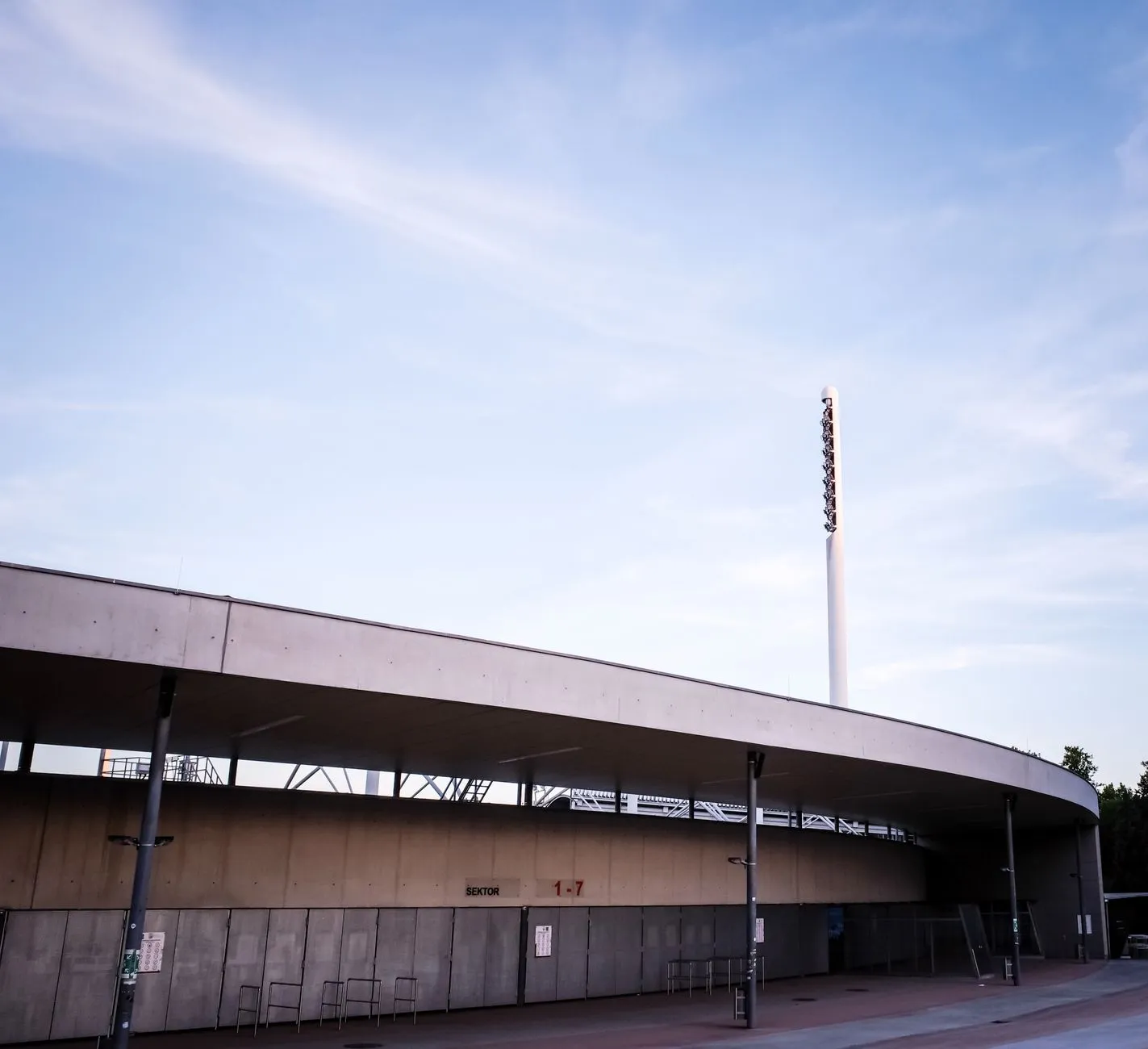 Zewnętrzna część stadionu z betonowym zadaszeniem i masztem oświetleniowym pod błękitnym niebem