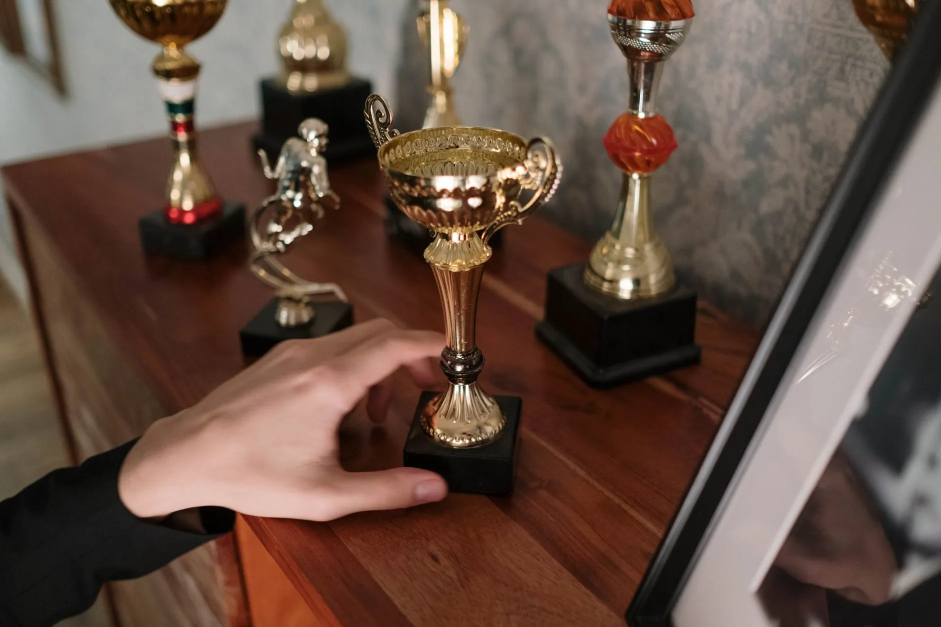 Dłoń sięgająca po złoty puchar stojący na drewnianej półce wśród innych trofeów