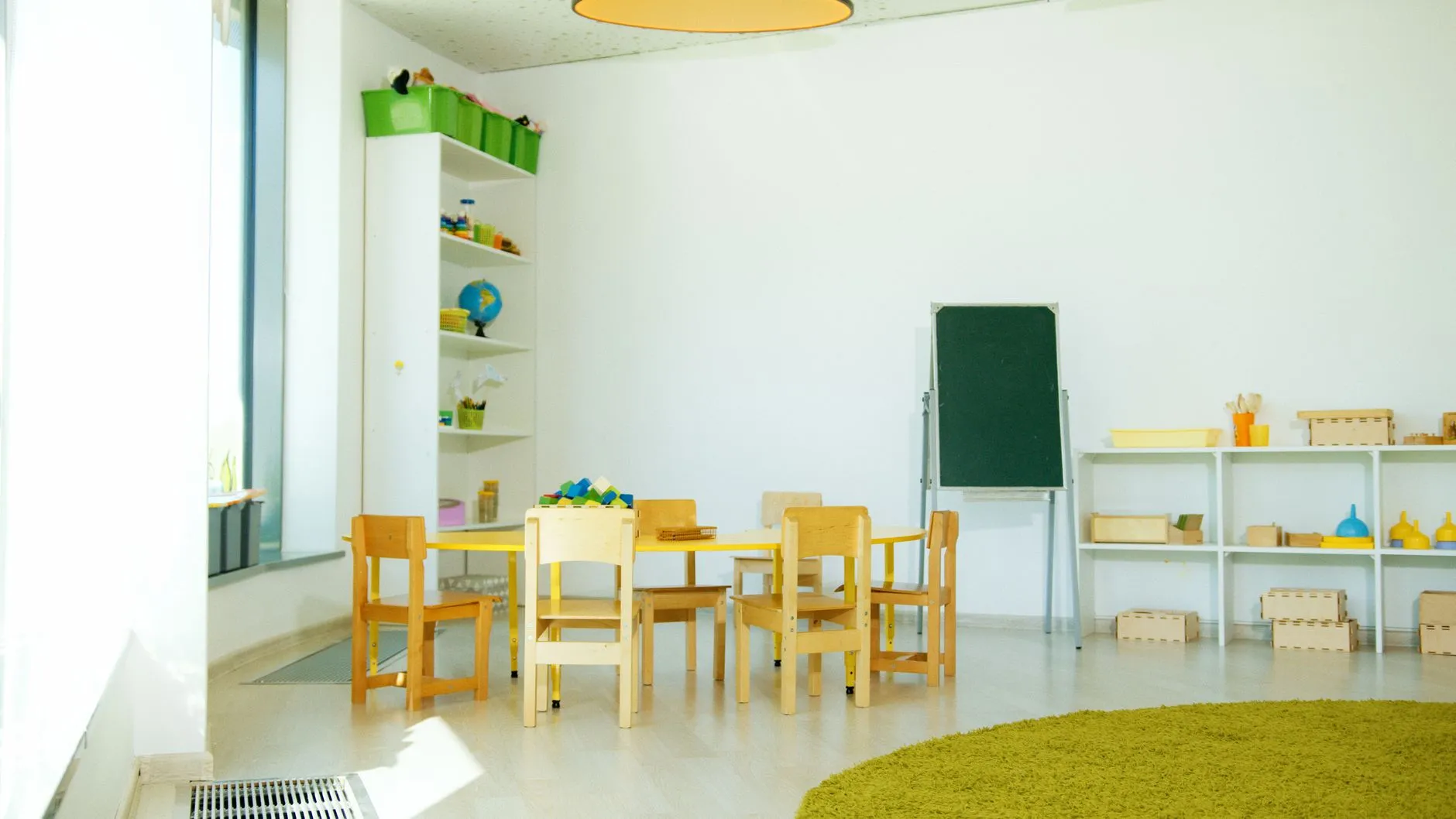 Jasna sala przedszkolna z drewnianymi krzesłami, stolikami, regałami i pomocami edukacyjnymi