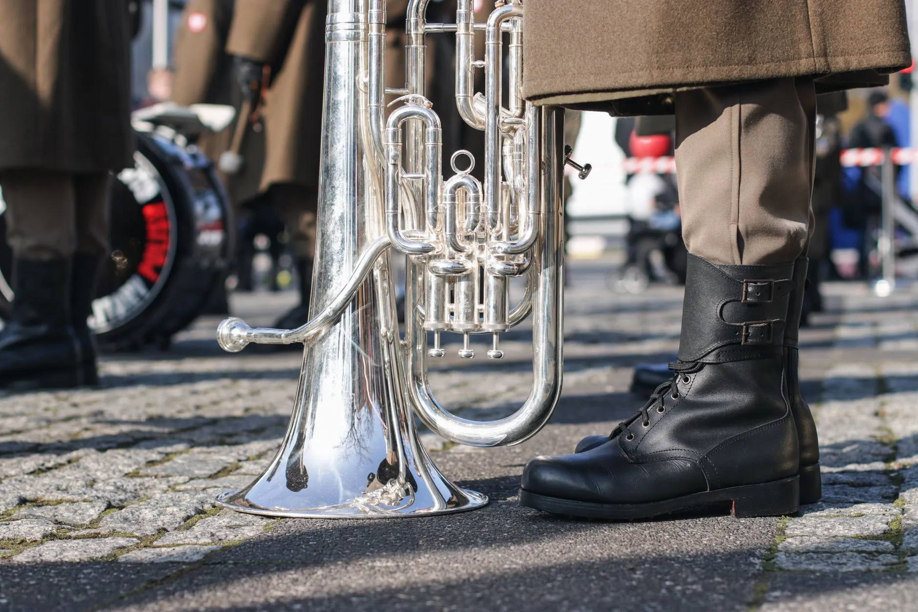 Srebrny instrument dęty obok czarnych sznurowanych butów osoby w mundurze na brukowanej ulicy