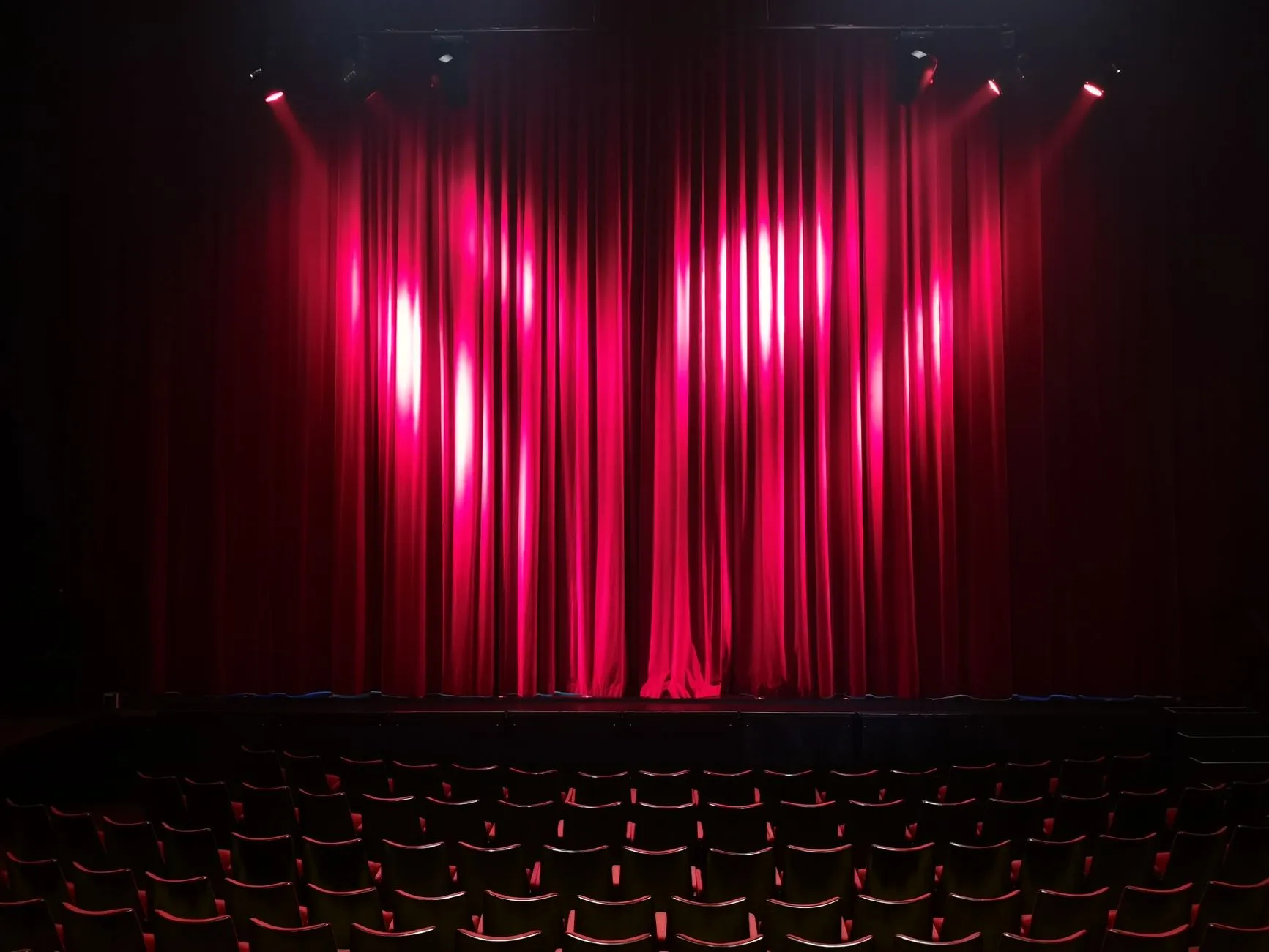 Czerwona kurtyna oświetlona reflektorami i rzędy pustych foteli w teatrze